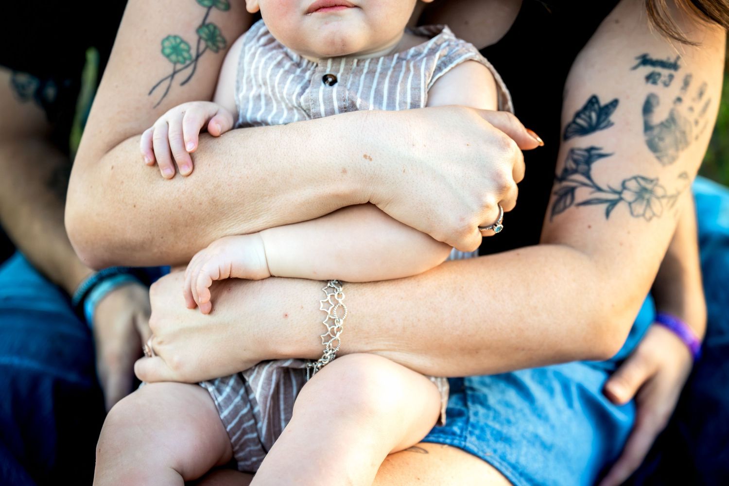 Two tattooed adults lovingly hold a baby in a striped outfit, showcasing a tender family moment outdoors.