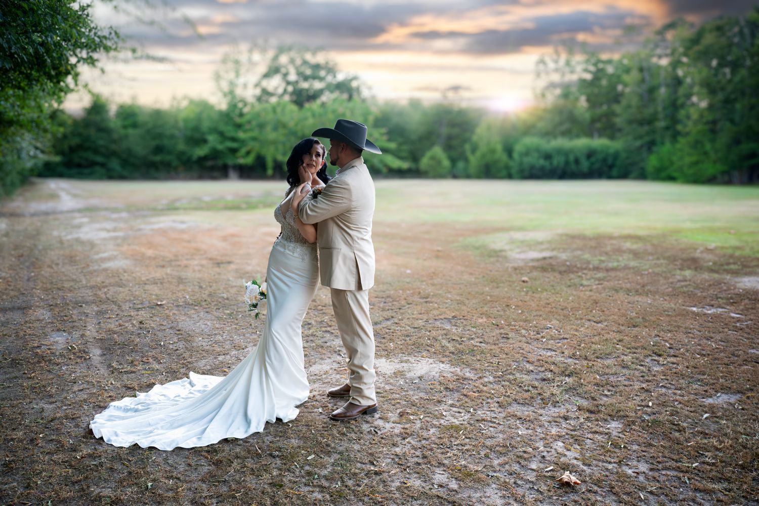 Bride and groom embrace at sunset on rustic grounds, bride in white gown, groom in tan suit with black hat.