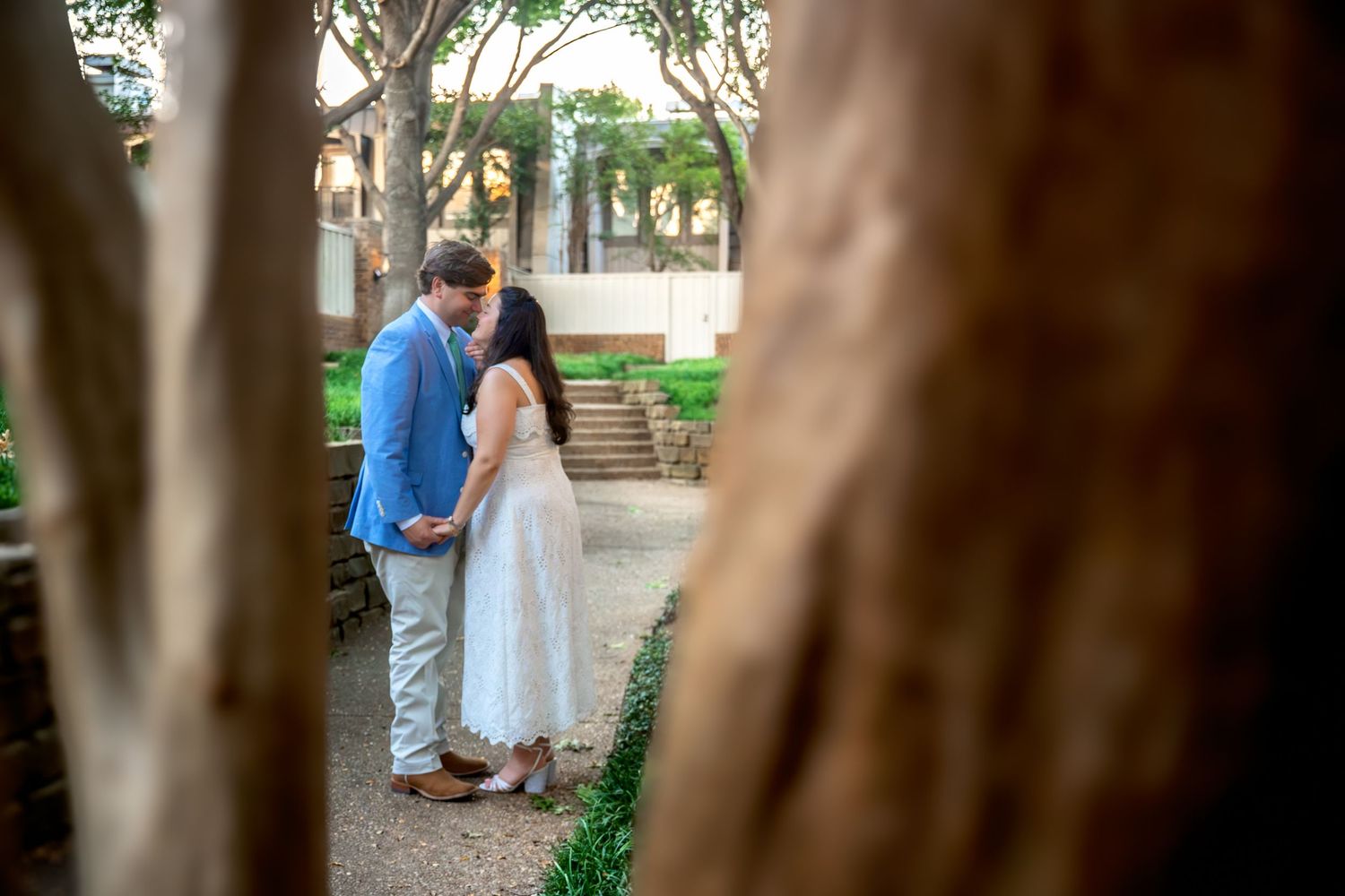 Couple sharing a romantic kiss outdoors among trees in a garden setting, man in blue suit and woman in white dress.