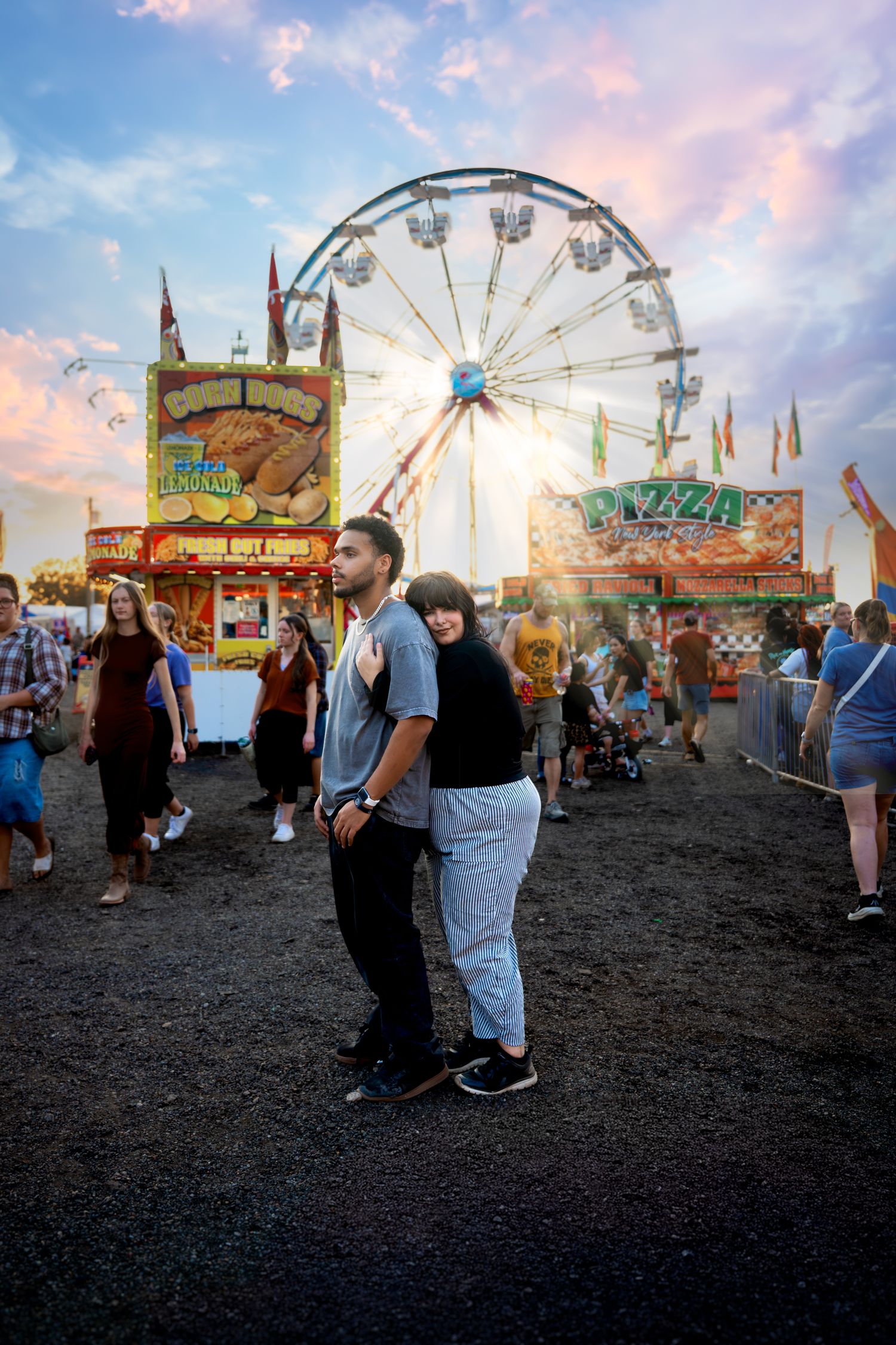 Couple standing together at a county fair with a Ferris wheel and food stands at sunset.