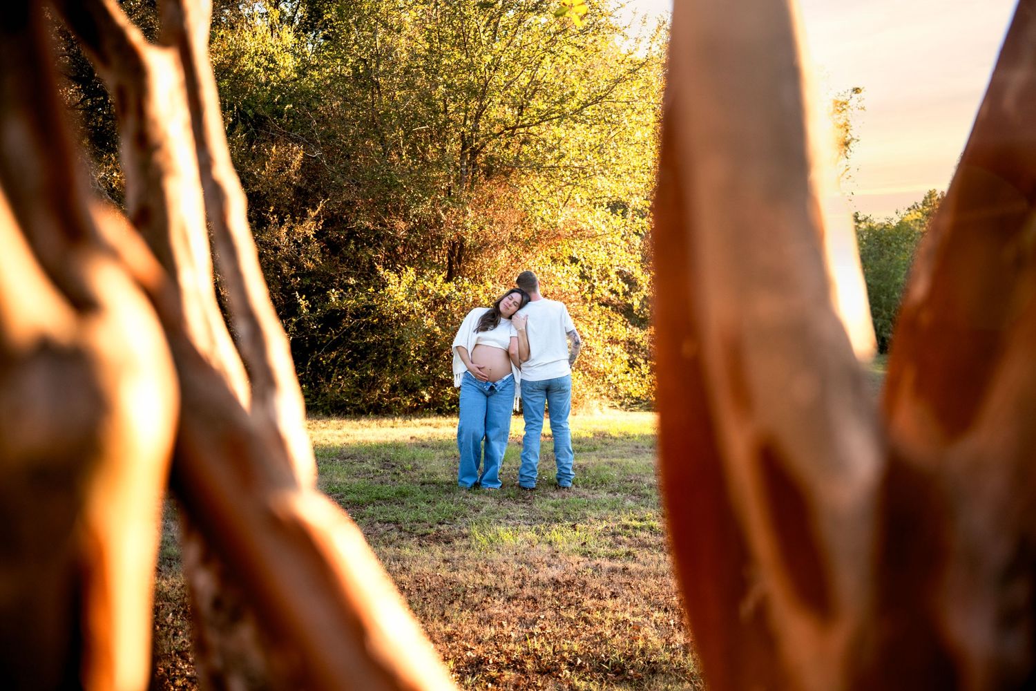 Couple embracing in a sunlit meadow framed by rustic tree trunks during a golden hour maternity photoshoot.