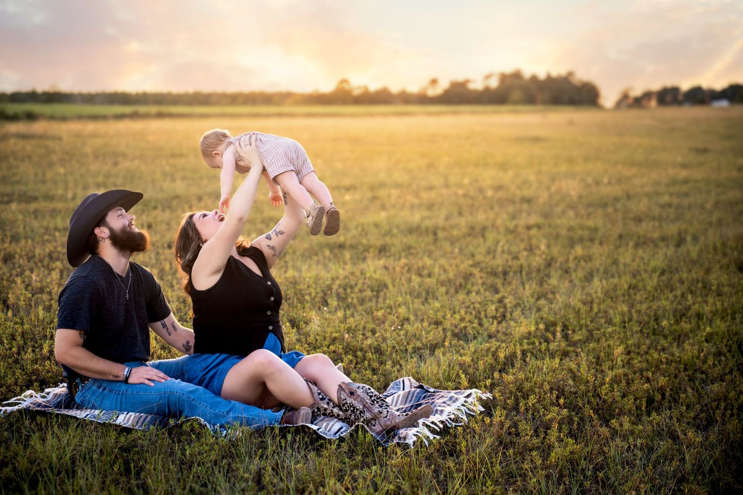 Family sitting on blanket in golden field at sunset, parents lifting baby in the air, enjoying outdoor picnic together.