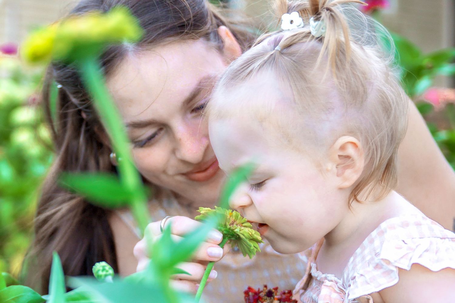Mother and baby girl smell flowers together in a lush green garden.