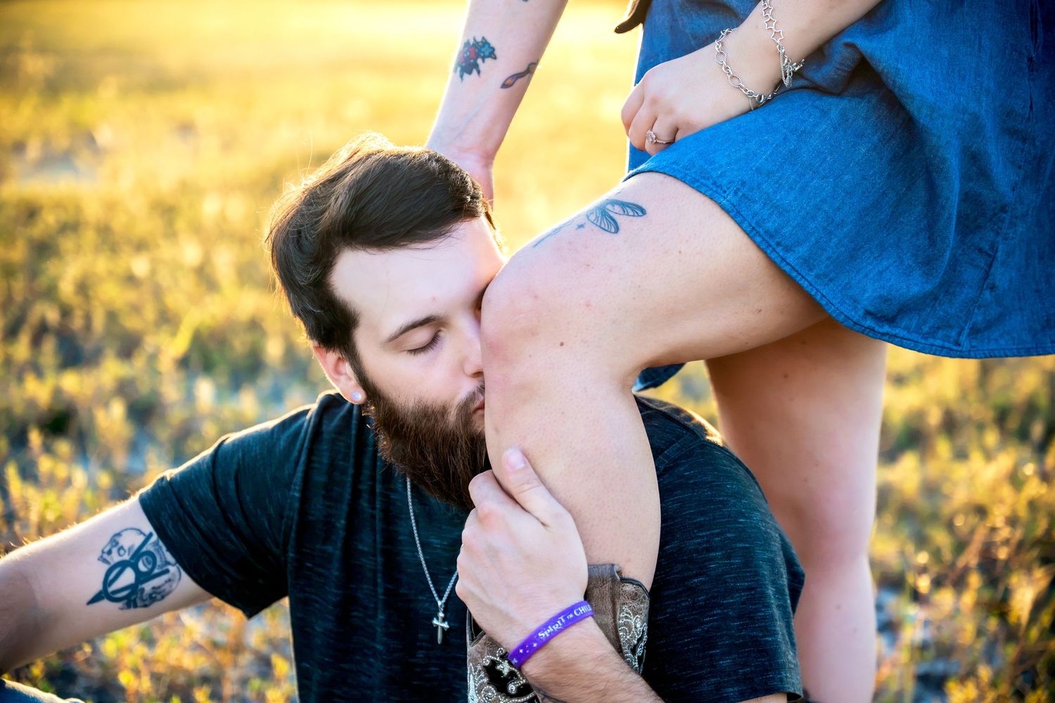 Bearded tattooed man kissing woman's hand in golden field at sunset.