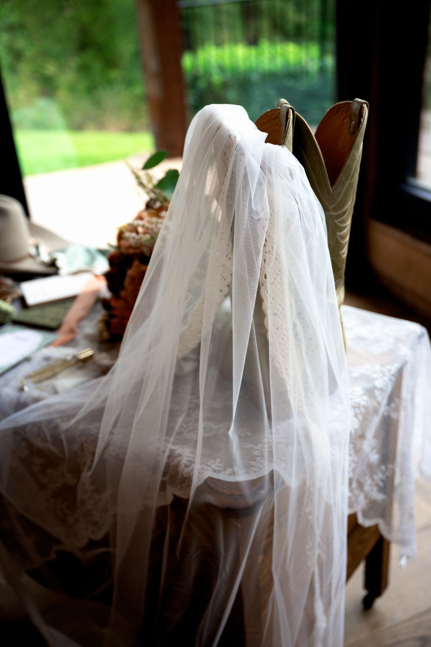 Bridal veil draped over cowboy boots on a rustic wooden chair near a window, perfect for a country wedding.