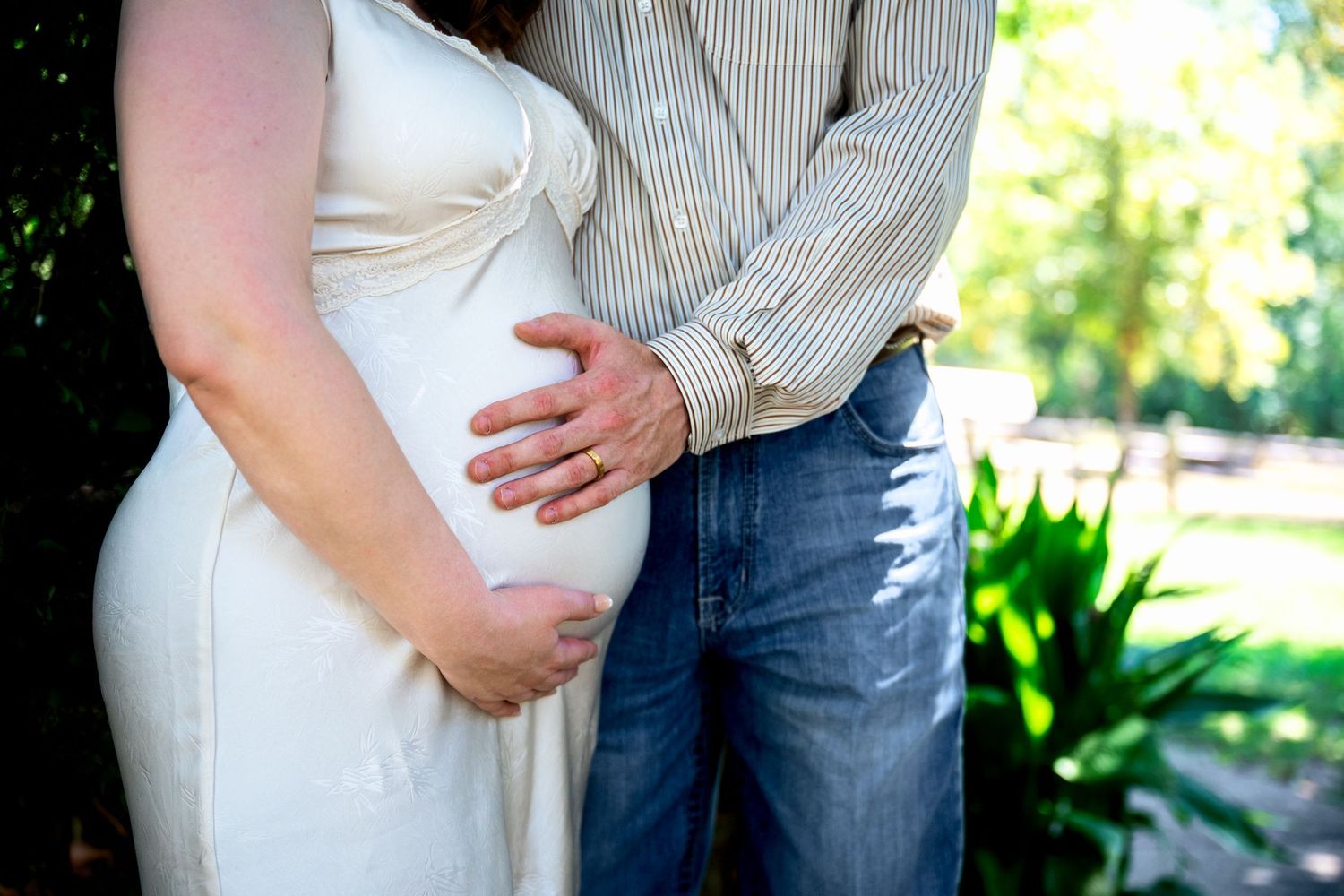 Pregnant woman in white dress with partner placing hand on baby bump outdoors in a maternity photo.