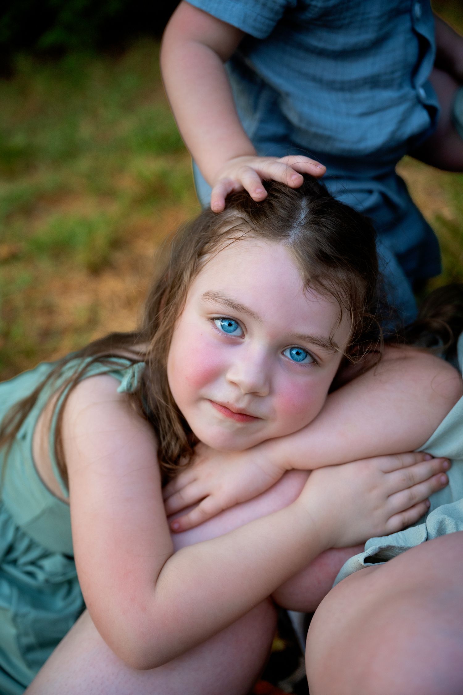 Sweet blue-eyed girl with arms crossed leaning against older sibling in outdoor portrait.