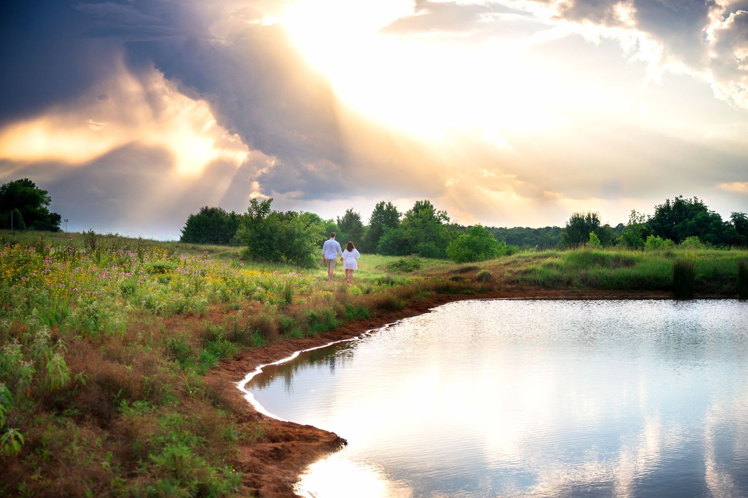 Scenic pond reflecting dramatic sunset rays through stormy clouds over lush green meadow with farmhouse in background.