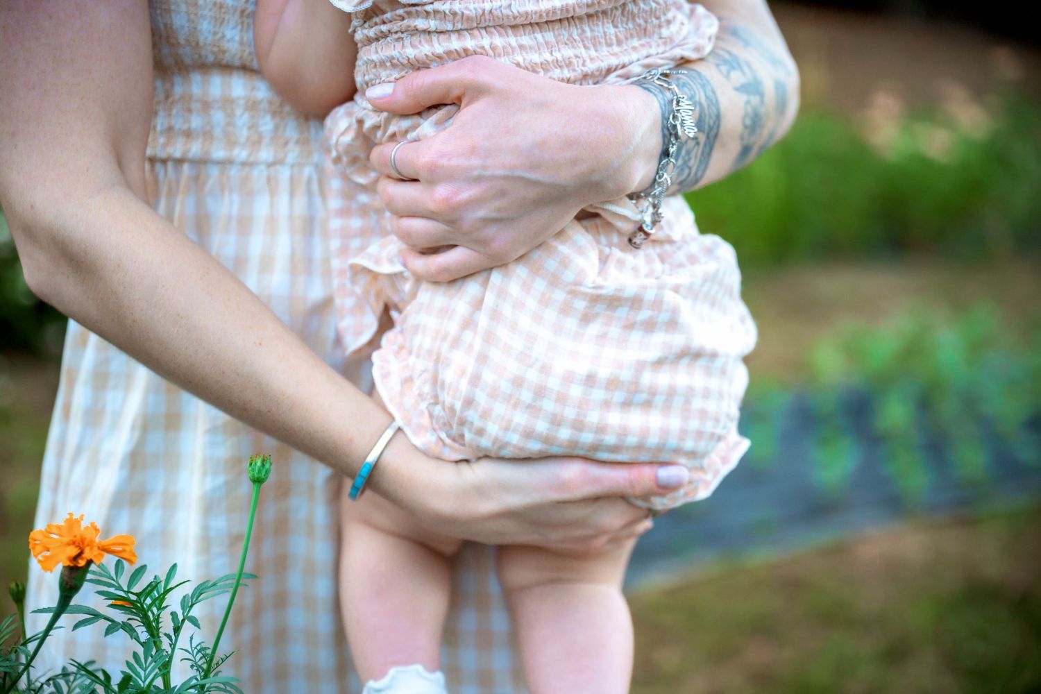Close-up of tattooed mother holding baby in gingham outfit near marigolds.