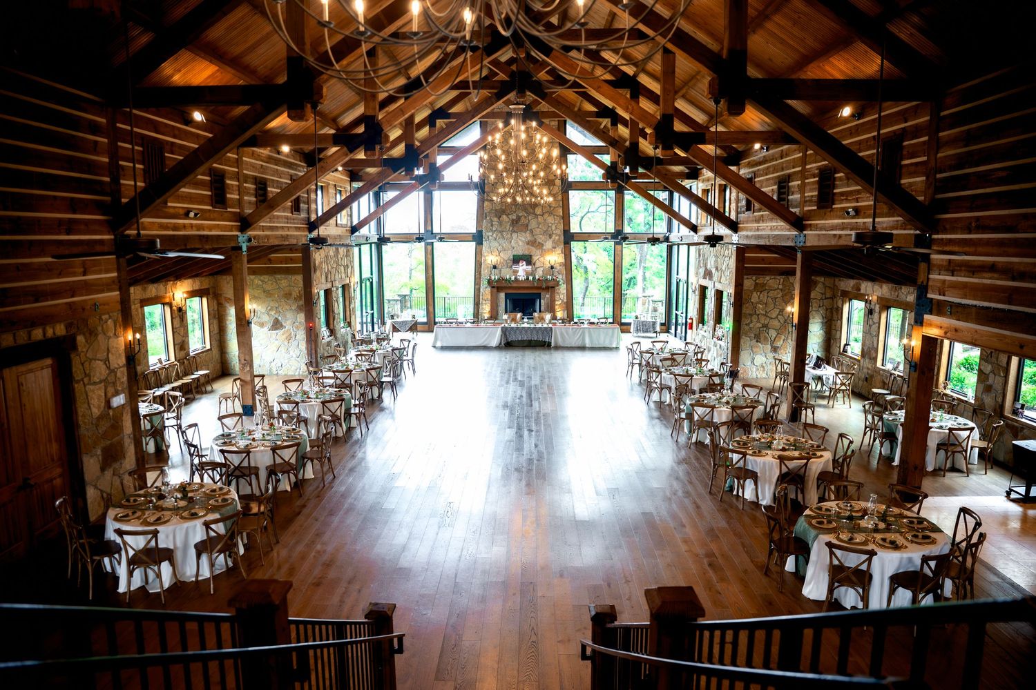 Rustic wedding venue with wood-beamed cathedral ceiling, chandelier, hardwood floors, and elegant round tables set for a reception.
