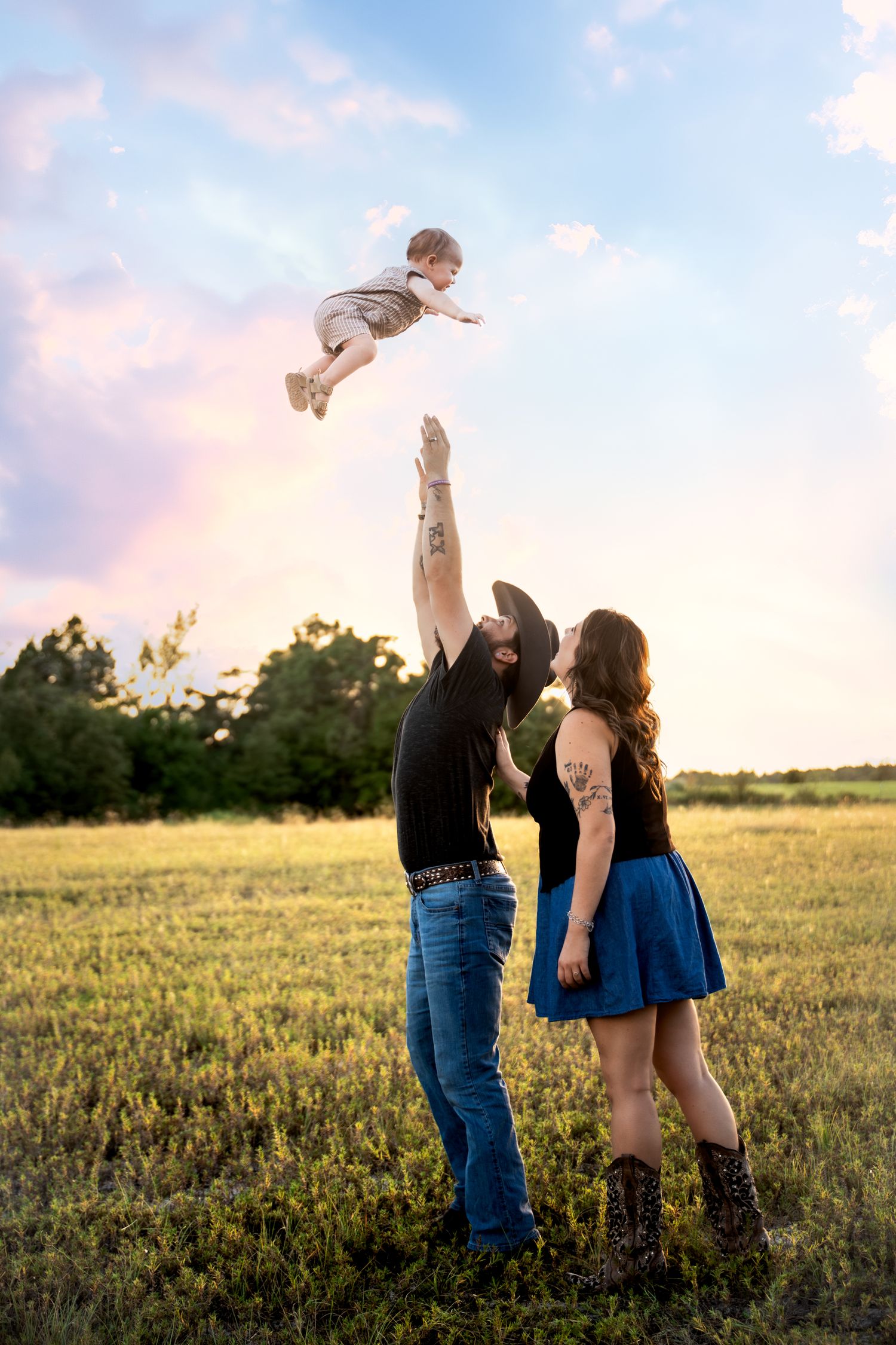A father tosses a toddler in the air while a mother watches in a green field at sunset.