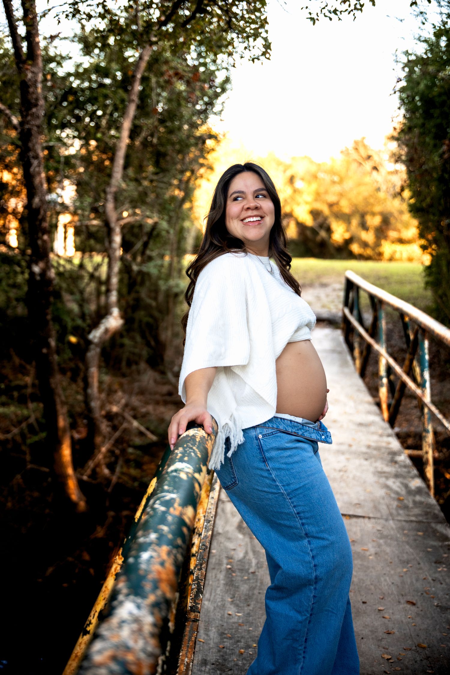 Smiling pregnant woman in white crop top and blue jeans leaning on a bridge railing outdoors at golden hour.