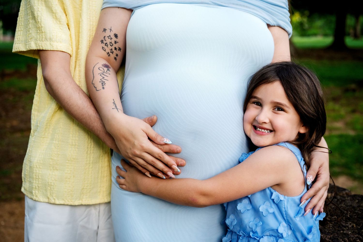 Young girl in blue dress hugging pregnant mothers belly outdoors in park setting.