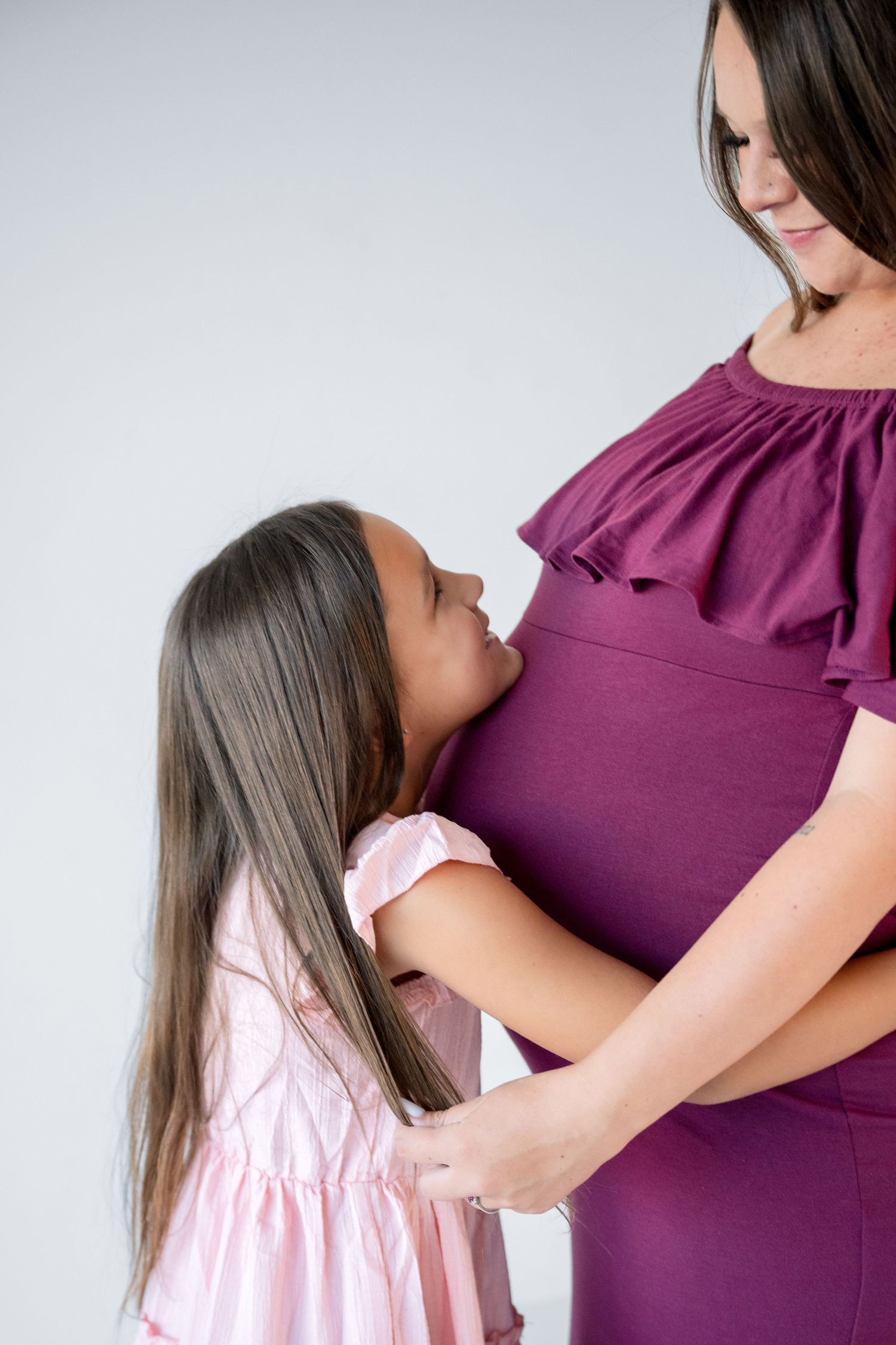 Young girl in pink dress looking up at pregnant mother in purple maternity dress.