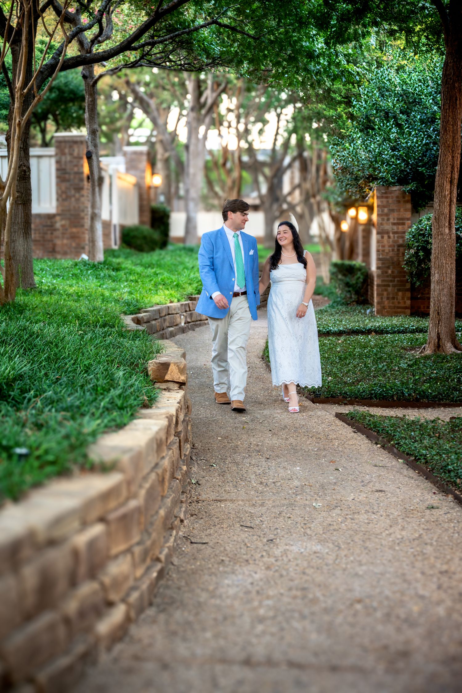 Couple walking hand in hand along a tree-lined garden path, man in blue blazer, woman in white dress.