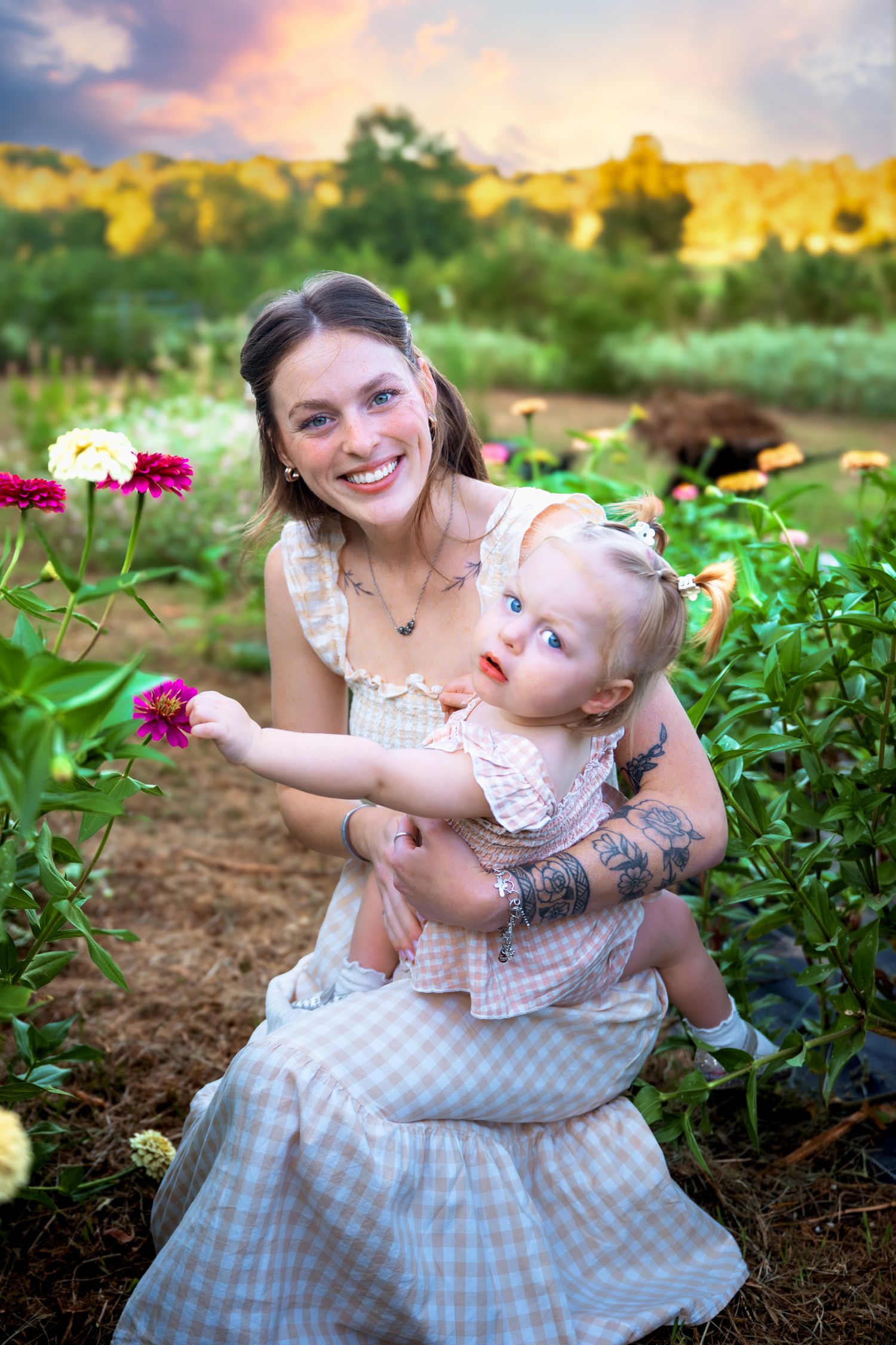 Smiling mother holds baby girl among colorful zinnia flowers in a garden at sunset.