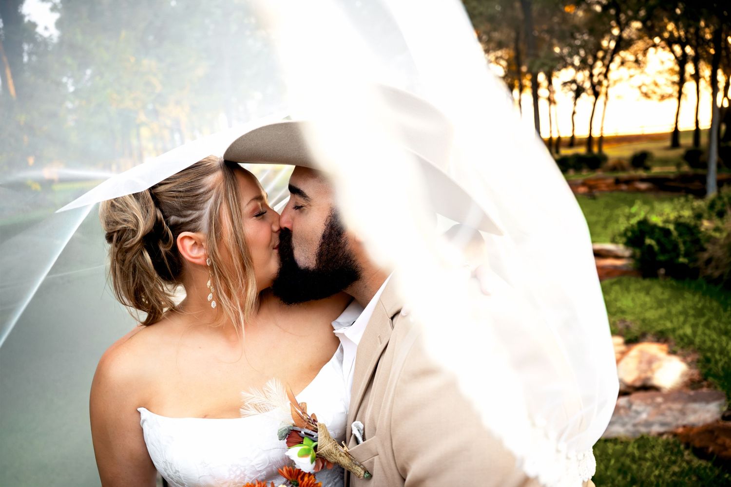 Bride and groom share a romantic kiss under a white veil at an outdoor wedding ceremony at sunset.