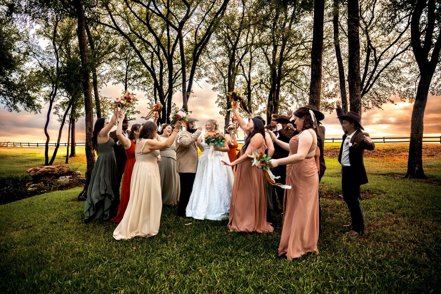 Wedding party celebrating outdoors at sunset, bride in white gown surrounded by bridesmaids in colorful dresses under trees.