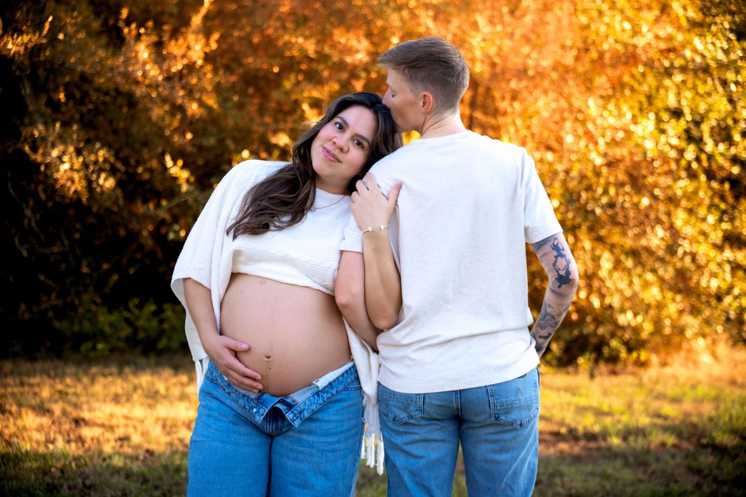 Pregnant woman in white top and jeans poses with partner outdoors during golden hour fall foliage maternity photo session.