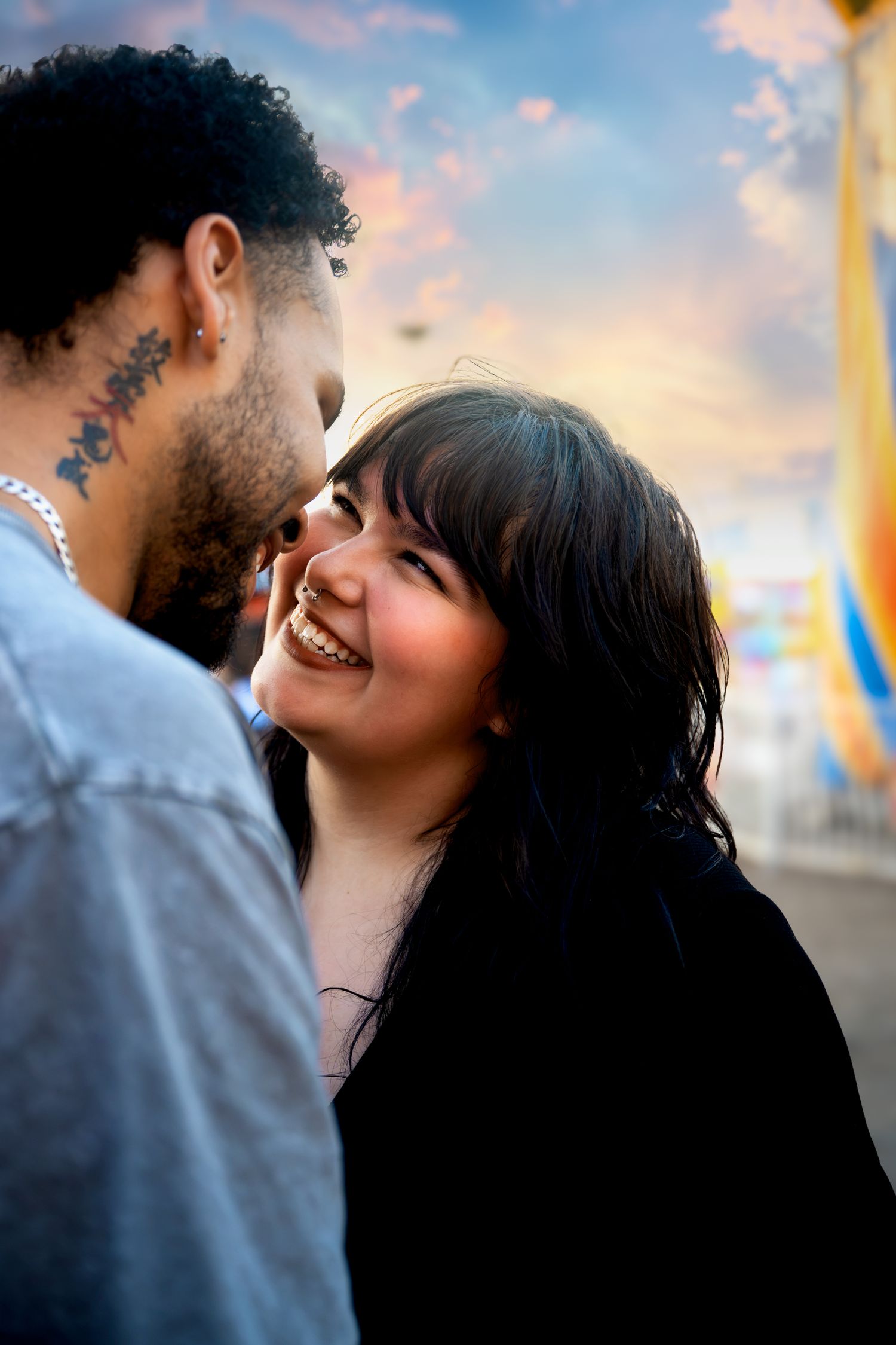 A smiling young woman gazes up at a man with neck tattoos at a colorful outdoor fair during golden hour.