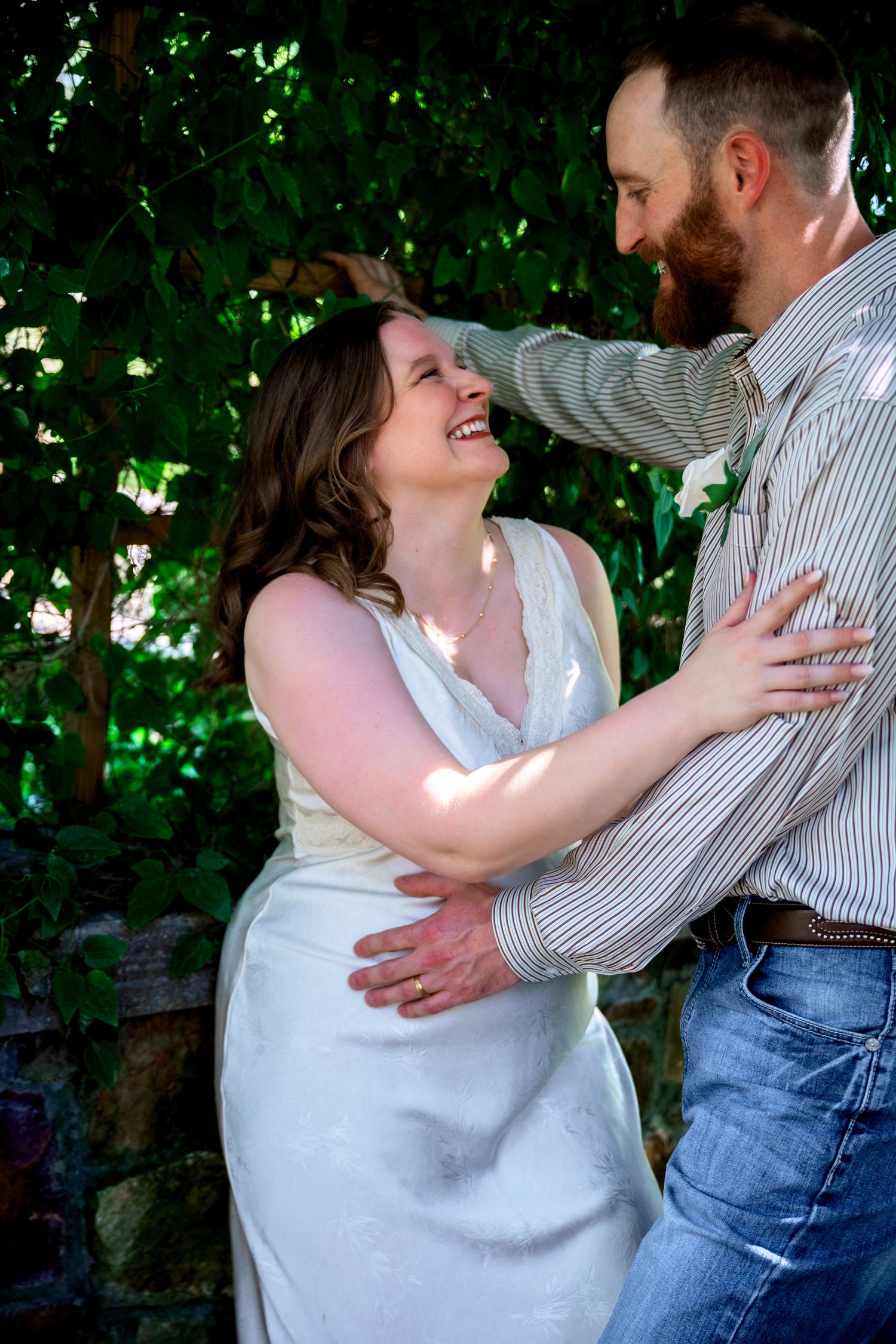 A joyful bride in white dress embraces groom in striped shirt outdoors, laughing together among green foliage.