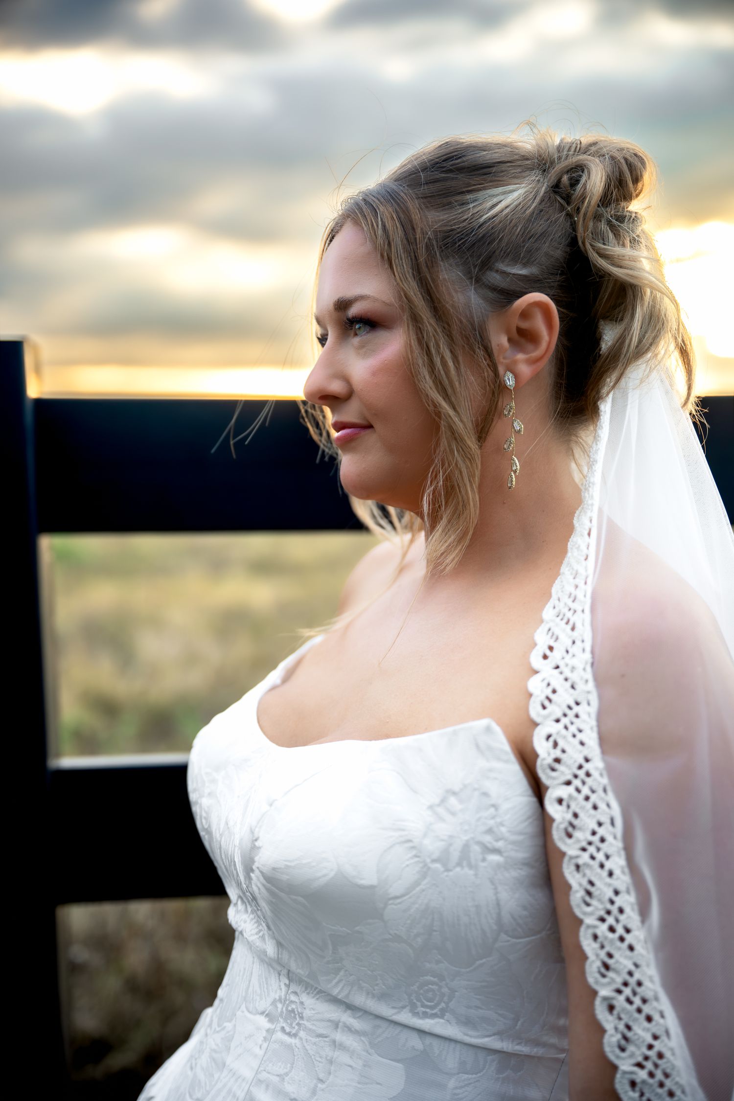 Bride in white lace gown and veil gazes sideways at sunset, wearing drop earrings, hair styled in updo.