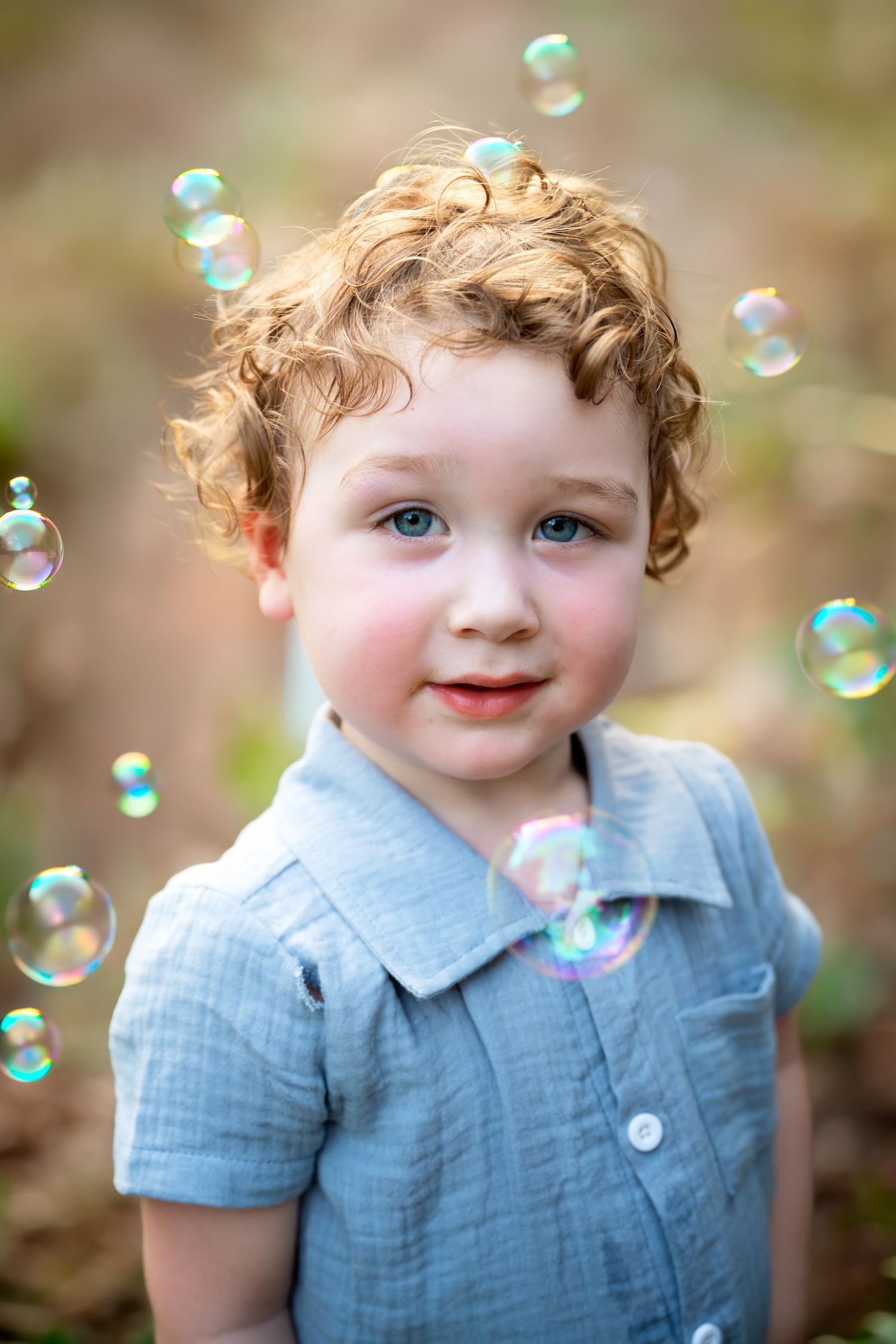 Adorable curly haired toddler boy with blue eyes surrounded by floating soap bubbles outdoors.