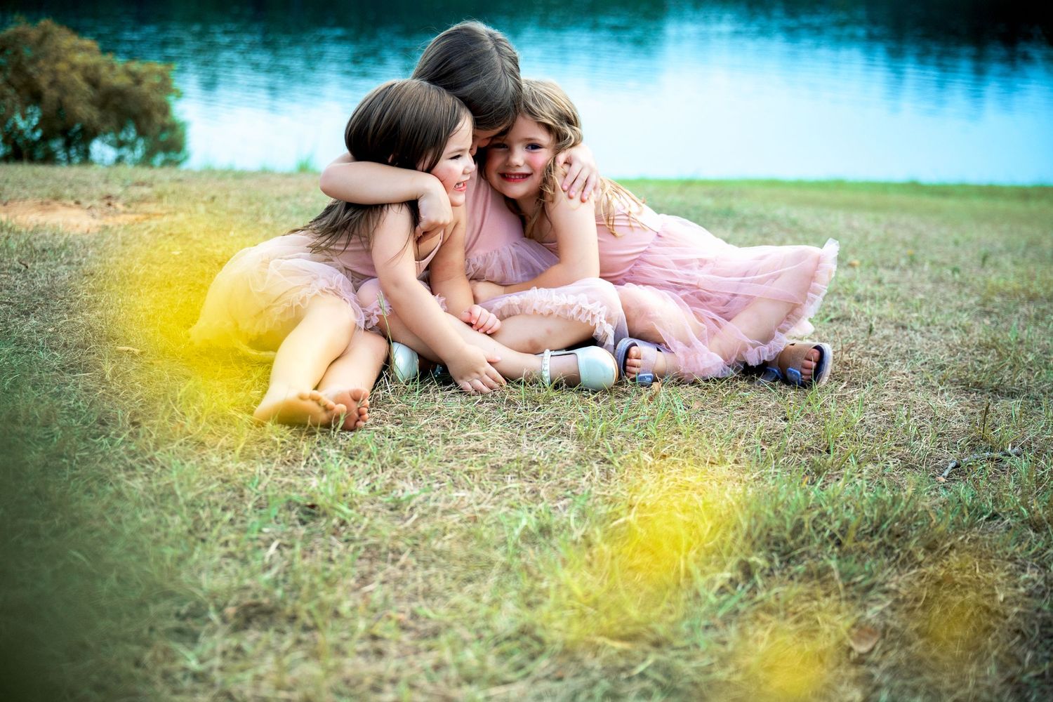 Three sisters in pink dresses hug and laugh together on grass by lake.