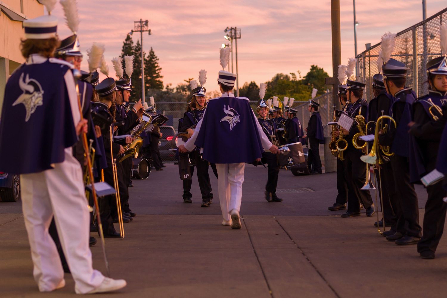 Marching band members line up along a walkway at sunset wearing white and navy uniforms while holding instruments.