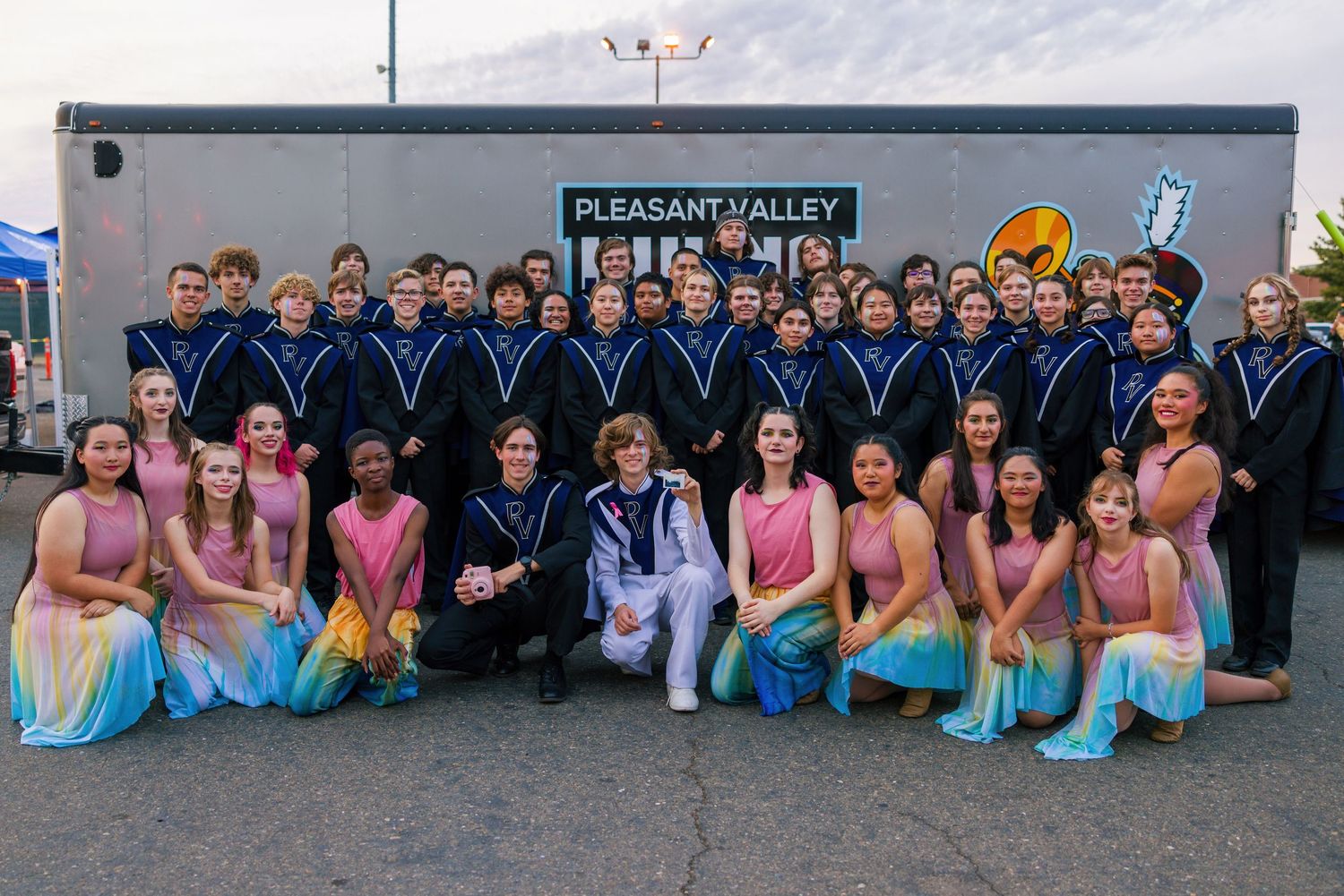 Band members in blue uniforms and performers in pastel dresses pose together in front of a trailer.