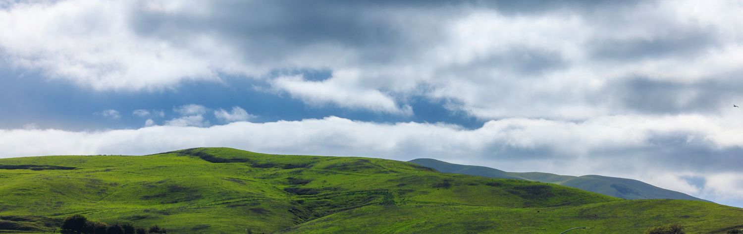 Rolling green hills stretch across a scenic mountain landscape under dramatic cloudy skies.