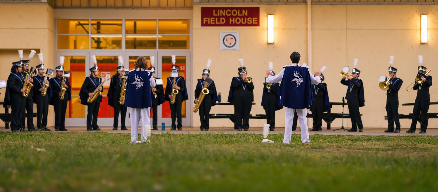 Band members in navy and white uniforms stand in formation on a field during an evening performance.