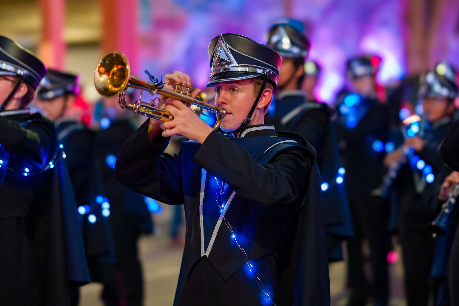 A trumpet player performs with a military band against a backdrop of blue stage lighting during a formal event.