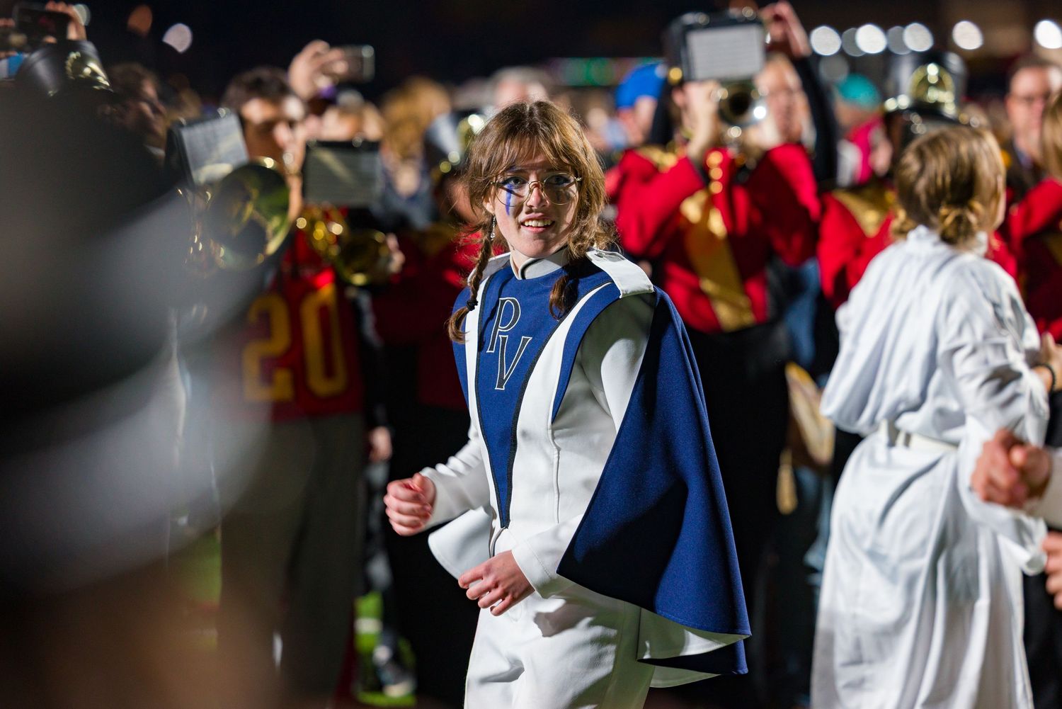 A performer in a blue and white cape walks among a crowd with red-uniformed band members in the background.