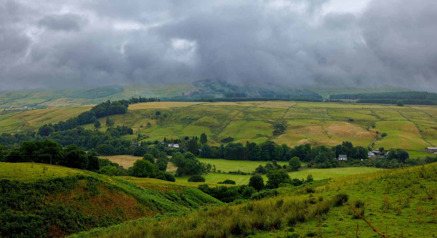 Rolling green hills and mountains under dramatic cloudy sky in panoramic landscape shot.