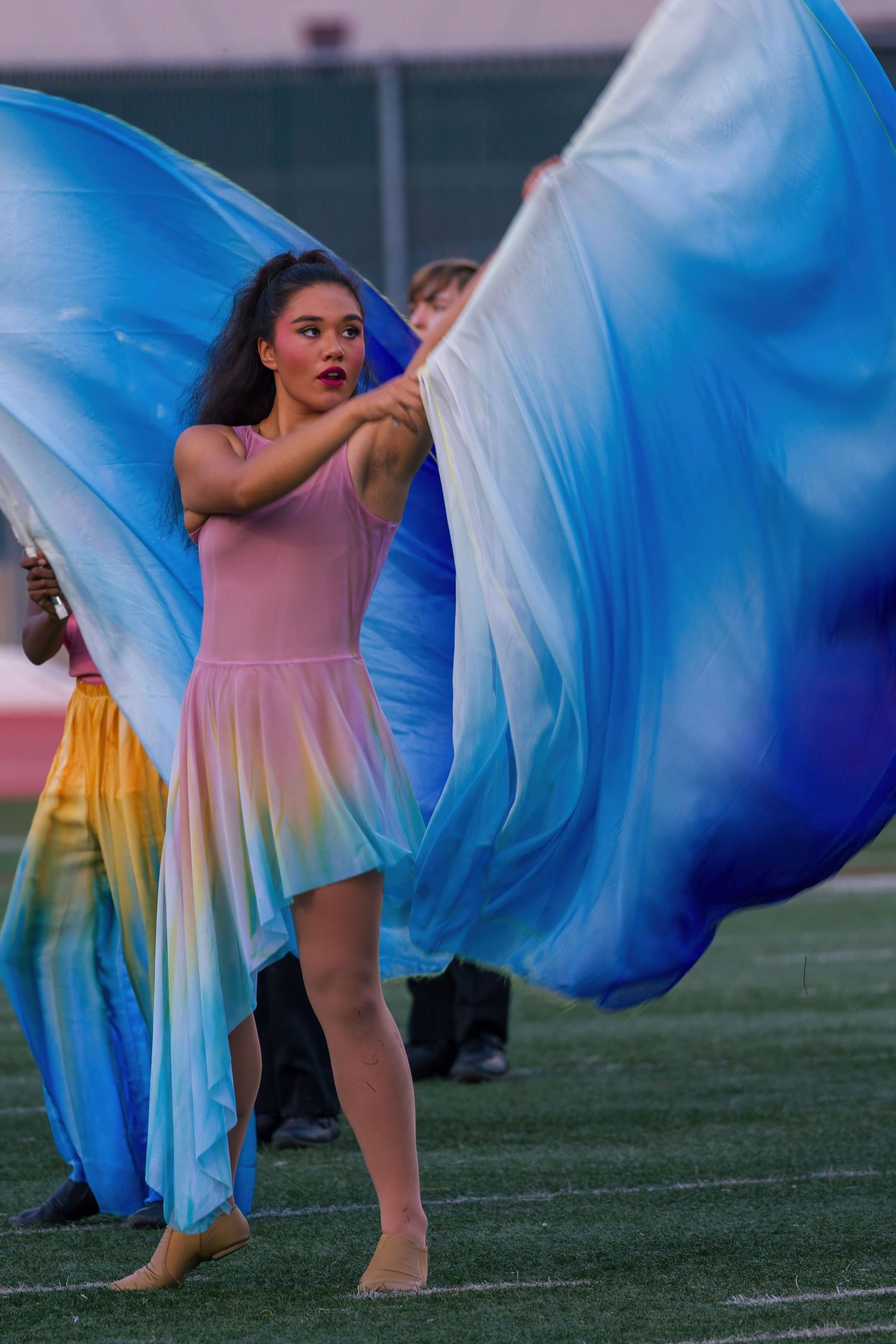 Color guard performers wave flowing blue and white flags while wearing pink and yellow dresses during a field show performance.