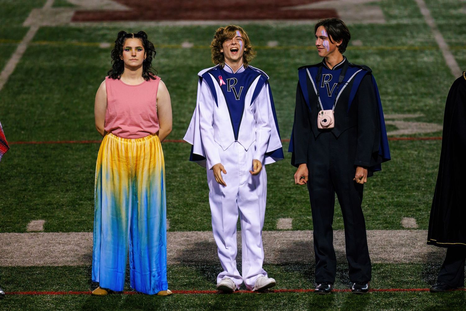 Performers in colorful costumes stand on a football field during a marching band performance.