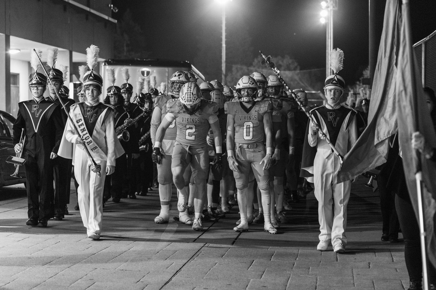 A group of marching band members in white uniforms perform during a nighttime parade under bright stadium lights.