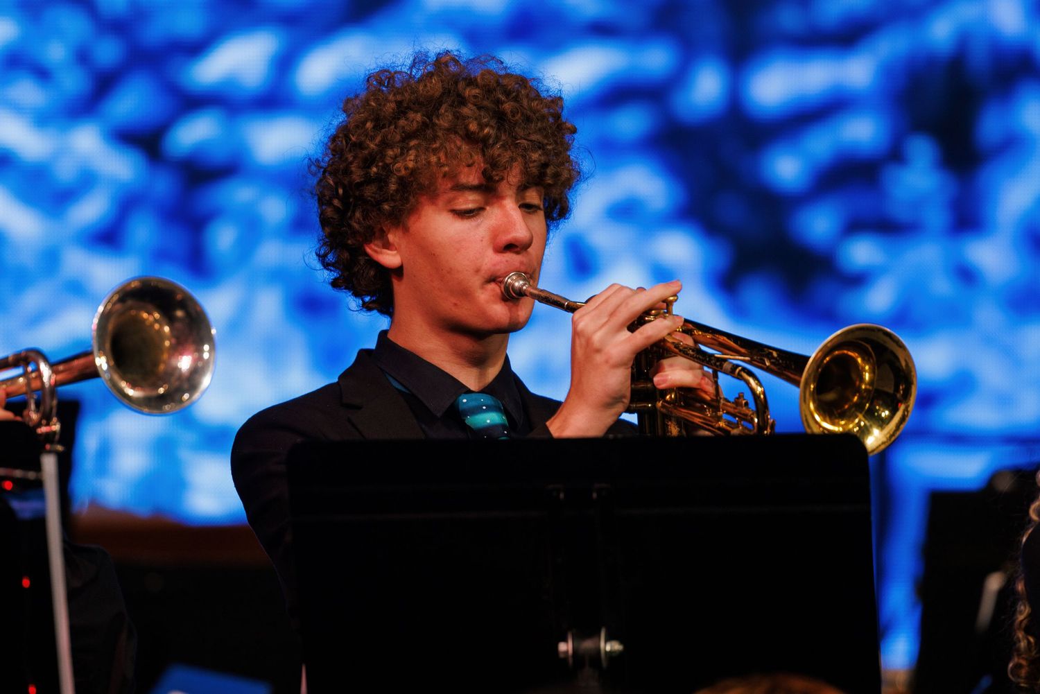 A trumpet player performing with multiple horns on stage against a bright blue abstract background.