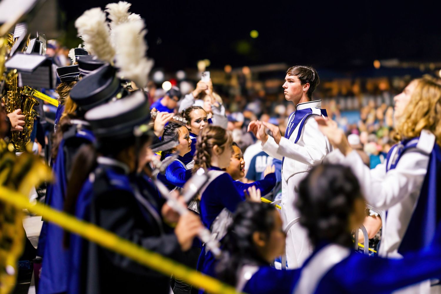 Marching band members perform from the bleachers during a nighttime game wearing blue and white uniforms.