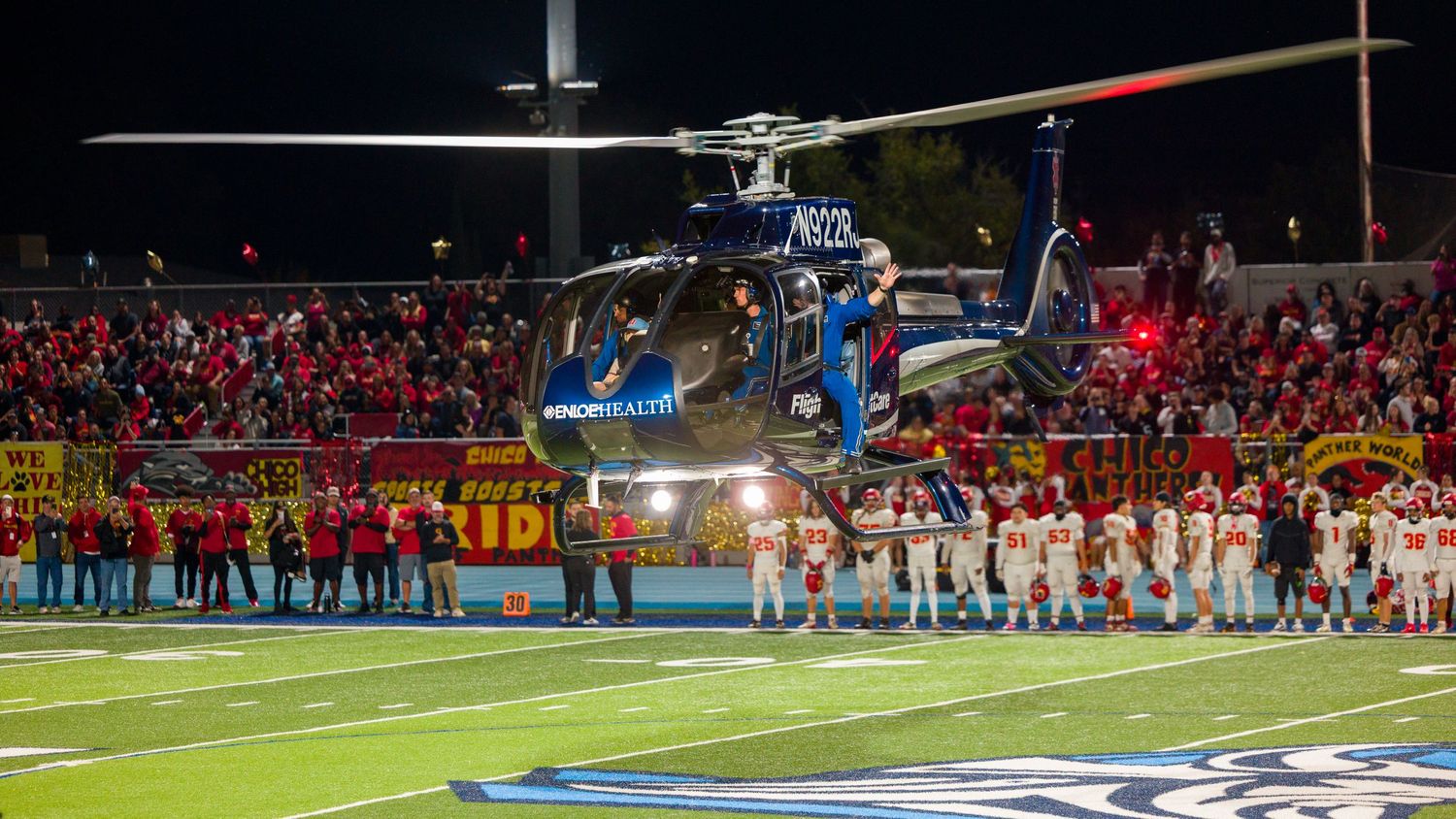 High school football players line up on the field during a night game under bright stadium lights.