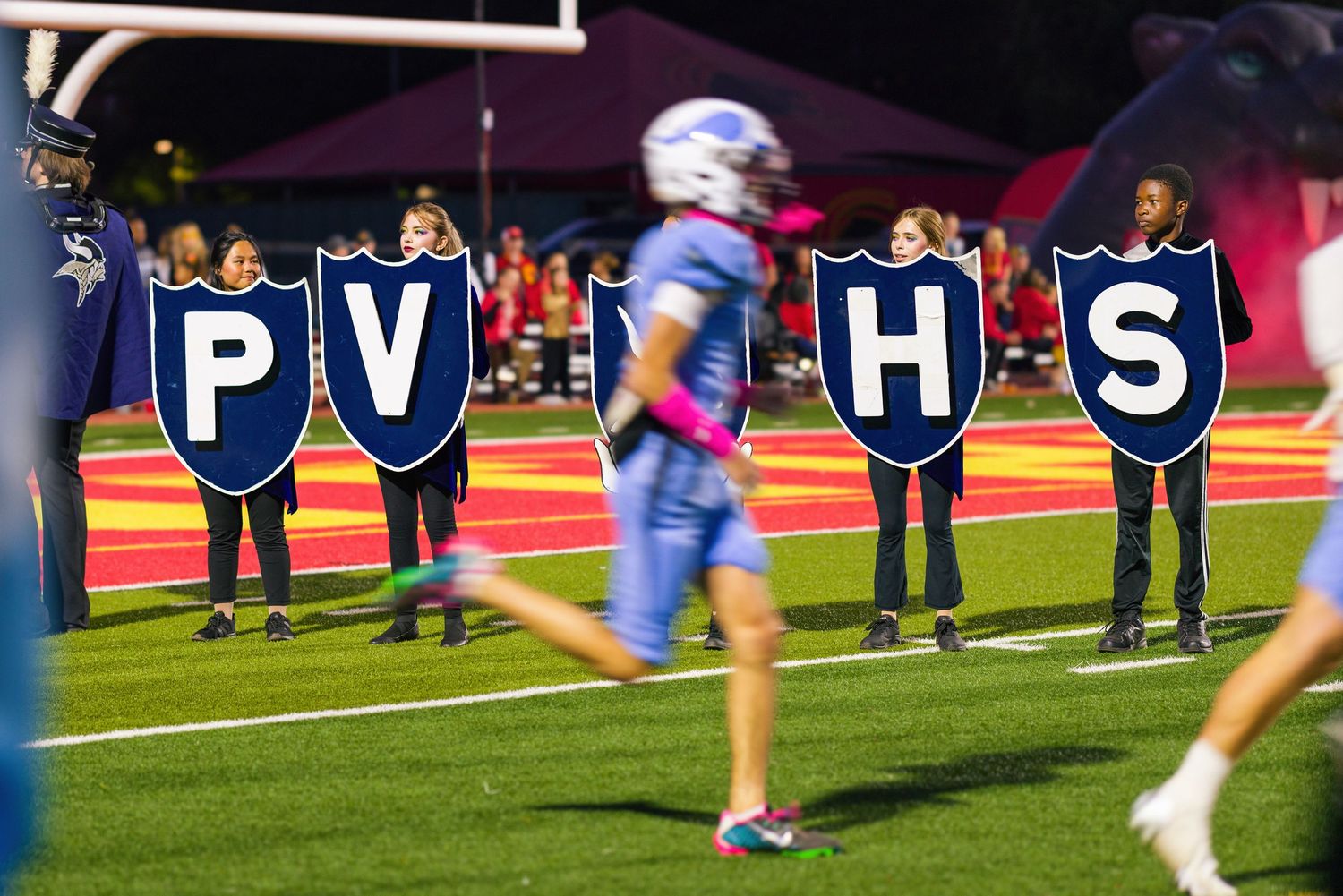 Students display letters spelling 'PV HS' during a nighttime football game on a colorful turf field.
