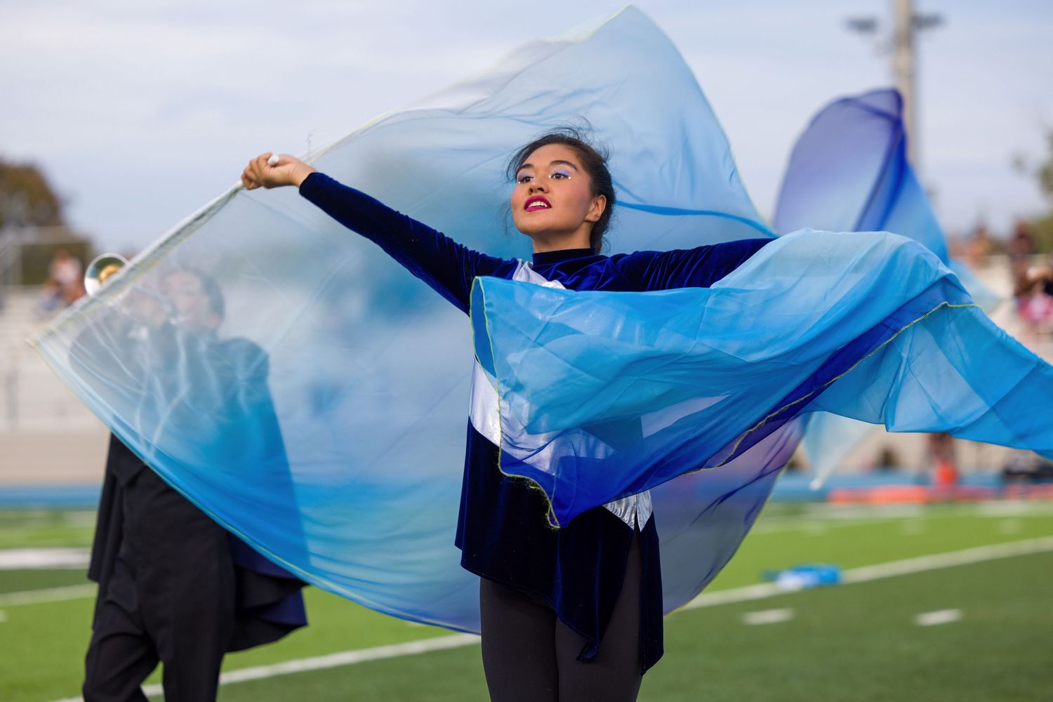 A color guard performer dances with flowing blue silk fabric on a football field during practice.