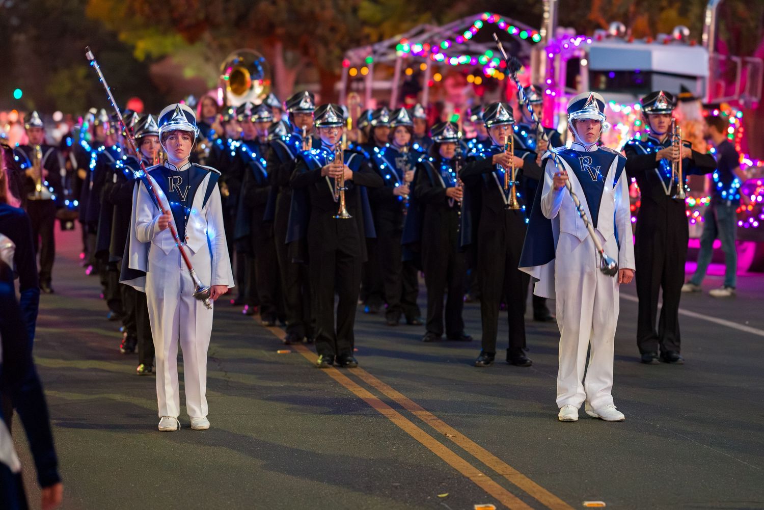 Marching band performers in white uniforms parade down a street at night with colorful lights illuminating the scene.