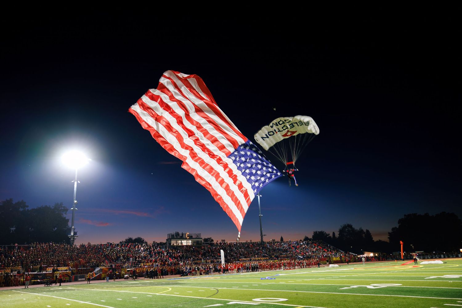 A parachutist descending with an American flag at a nighttime football stadium with floodlights illuminating the field.