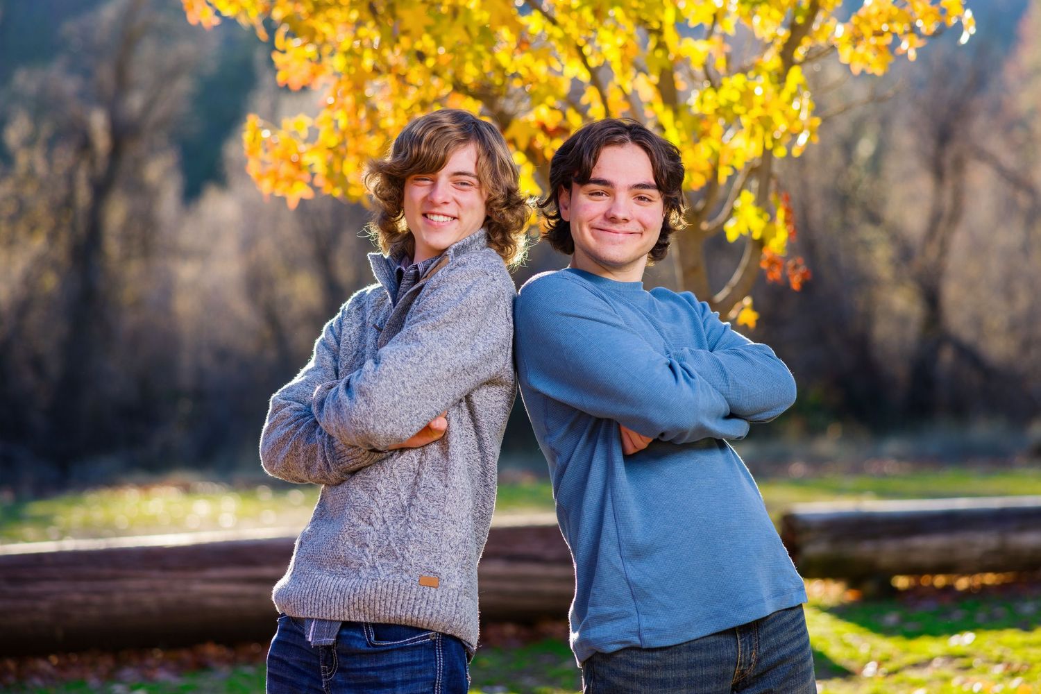Two people stand back to back smiling outdoors with autumn yellow leaves and foliage in the background.