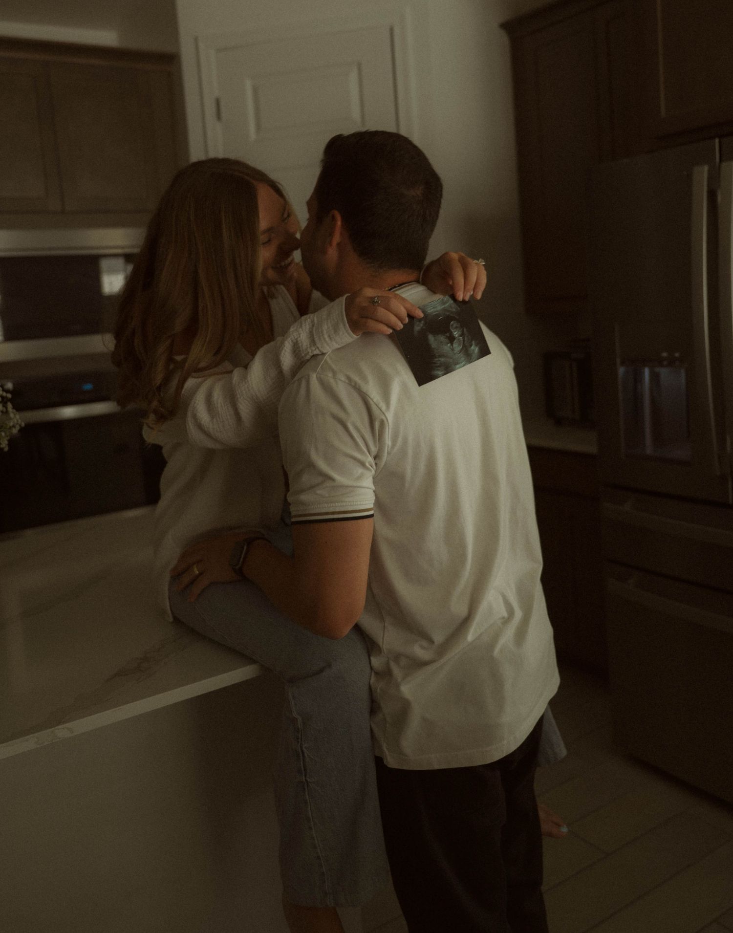 Couple sharing a kiss in a kitchen while holding an ultrasound photo, celebrating a pregnancy announcement.