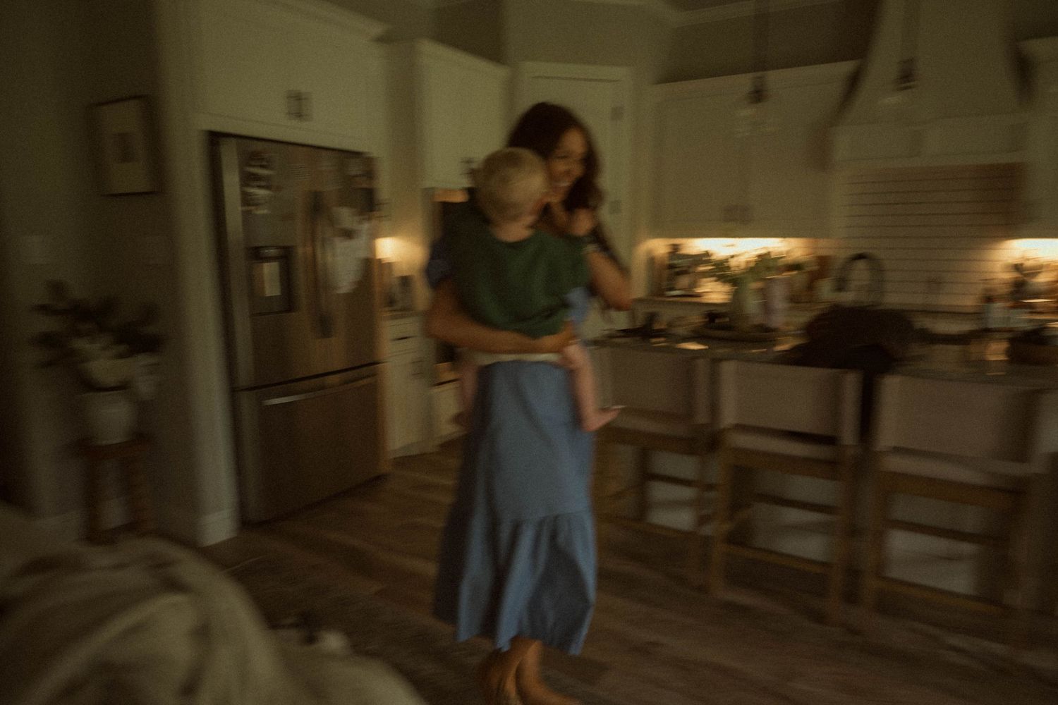 Woman holding a child in a dimly lit modern kitchen with stainless steel appliances and bar stools.
