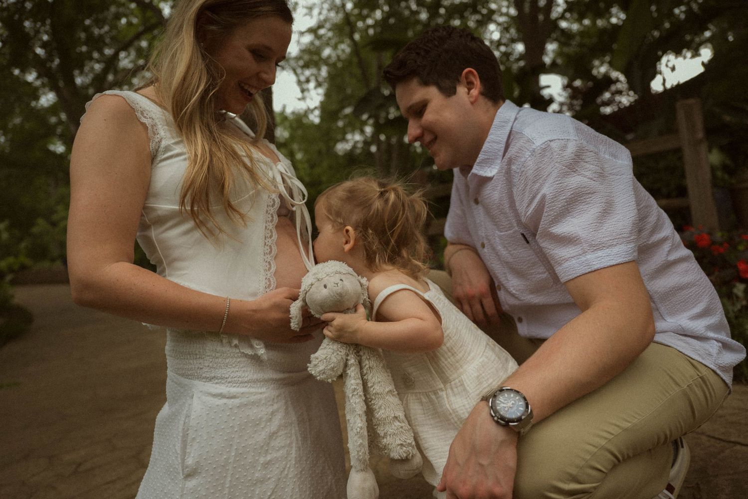 A young family with a toddler girl holding a stuffed bunny, sharing a tender moment outdoors in a garden setting.