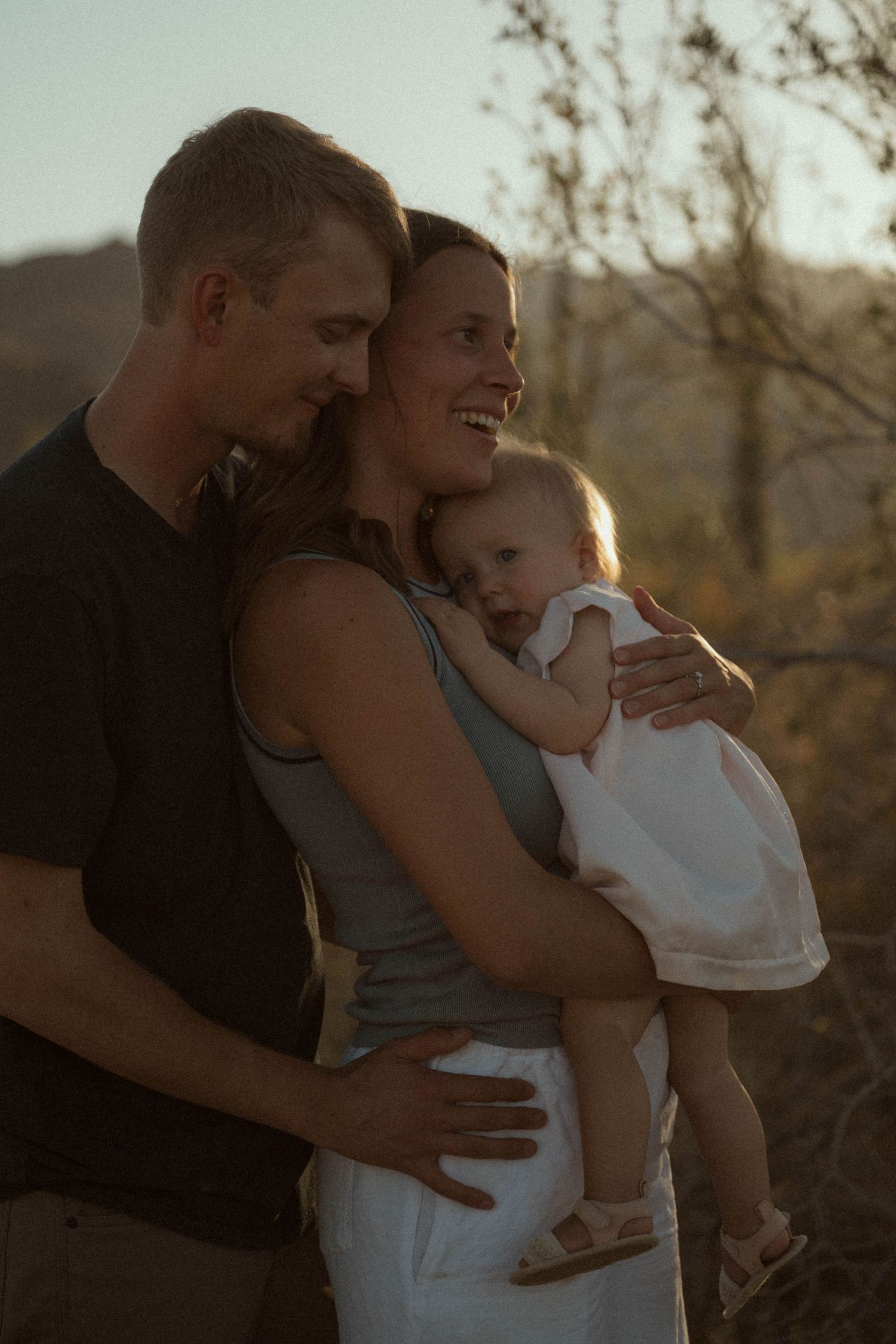 A happy family of three poses outdoors at golden hour, parents embracing while holding their baby girl.